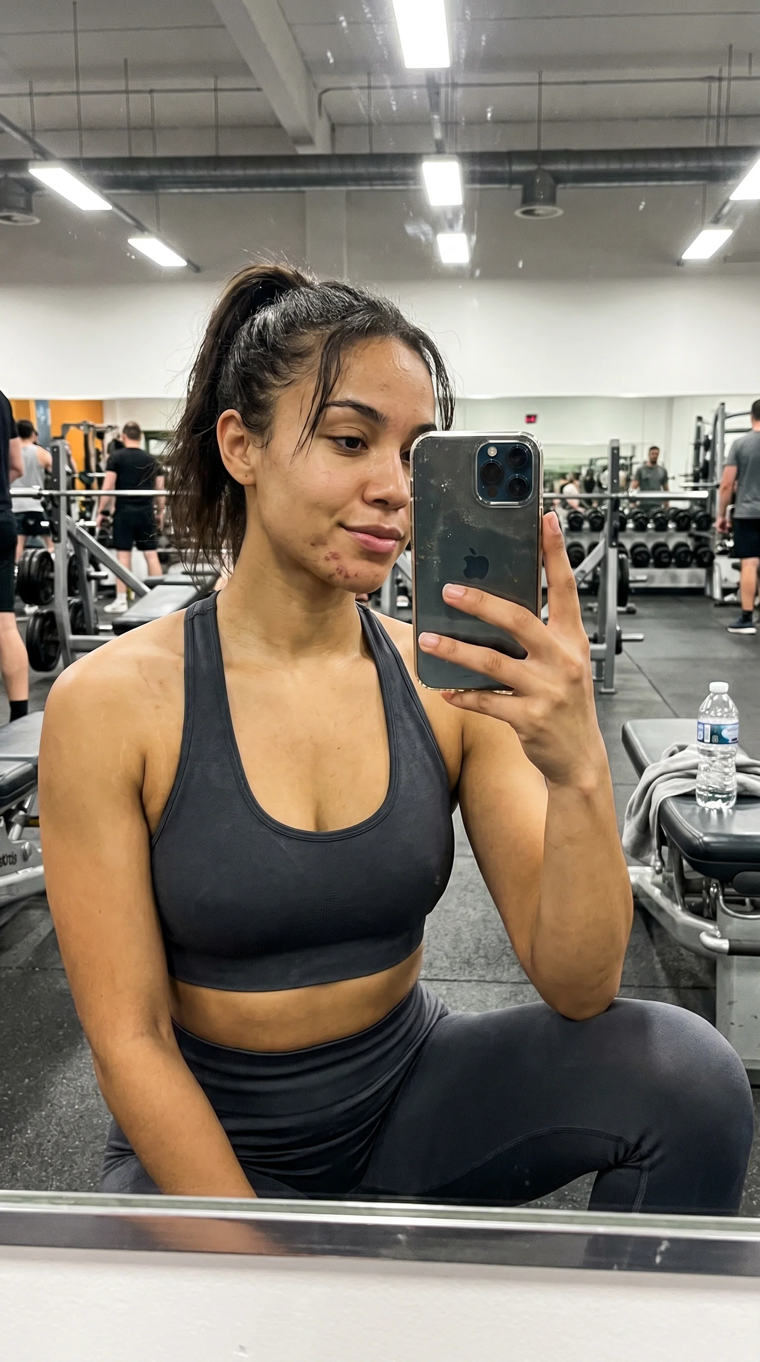 Selfie de academia: top esportivo escuro, academia real ao fundo com gente treinando, marcas de acne no queixo, espelho com dedadas.