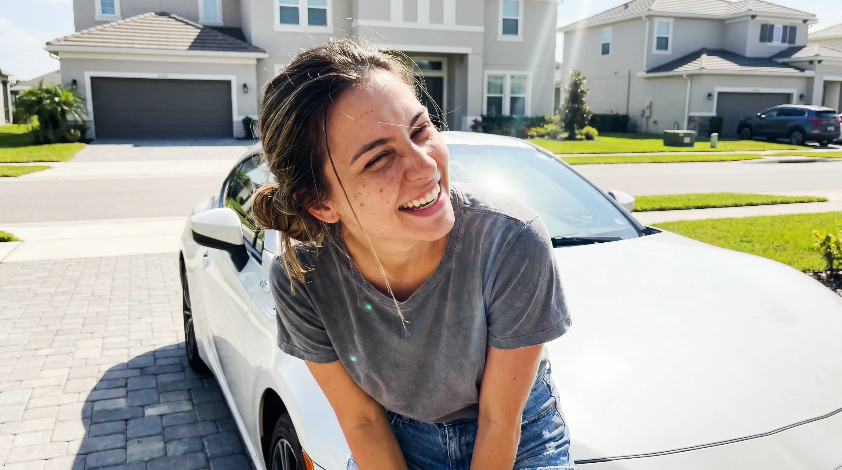 Mulher de coque baixo bagunçado apertando os olhos e rindo contra o sol forte de meio-dia, encostada num carro esportivo branco numa rua de condomínio, highlights estourados na lataria.