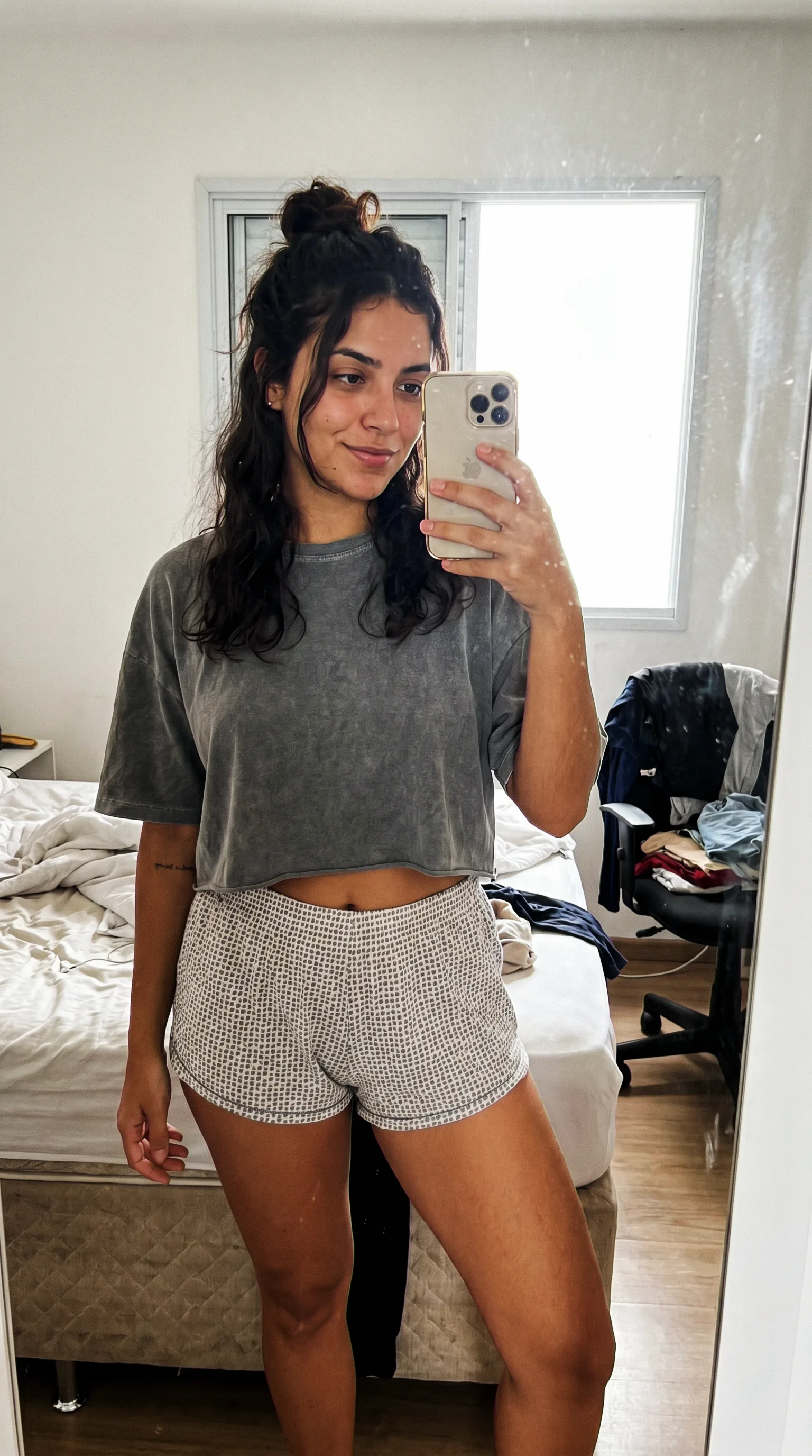 Selfie de espelho no quarto: camiseta cinza lavada, quarto real com cama box desfeita e pilha de roupa na cadeira, janela estourada de luz. Parece foto de celular, não imagem gerada.