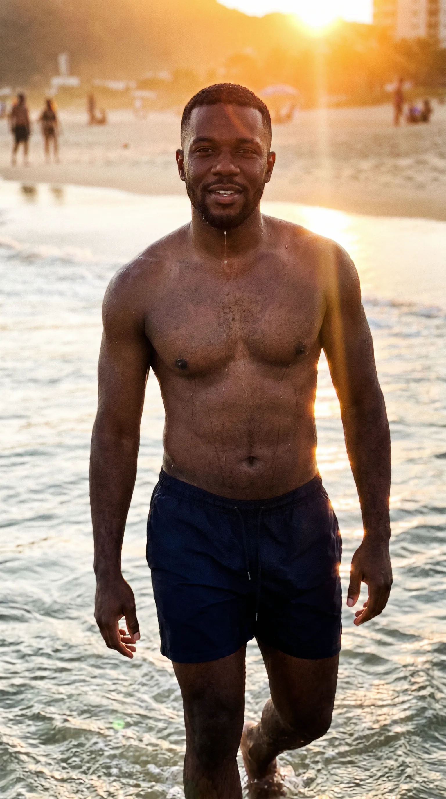 Homem negro de uns 38 anos saindo do mar no fim de tarde, água escorrendo, contraluz quente com flare, banhistas desfocados ao fundo.