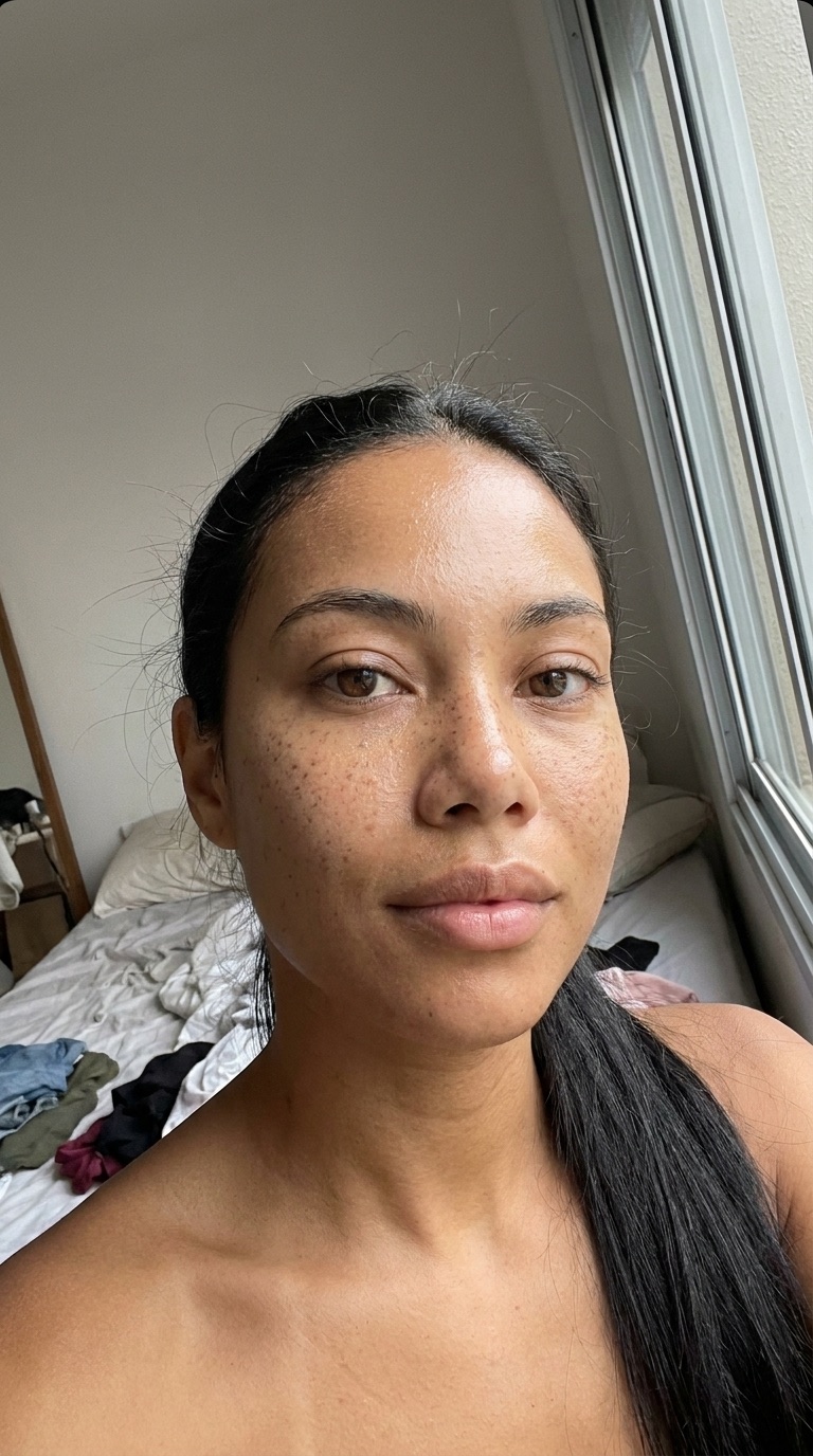 A Lia como selfie realista: poros visíveis, oleosidade na testa, quarto com luz de janela.